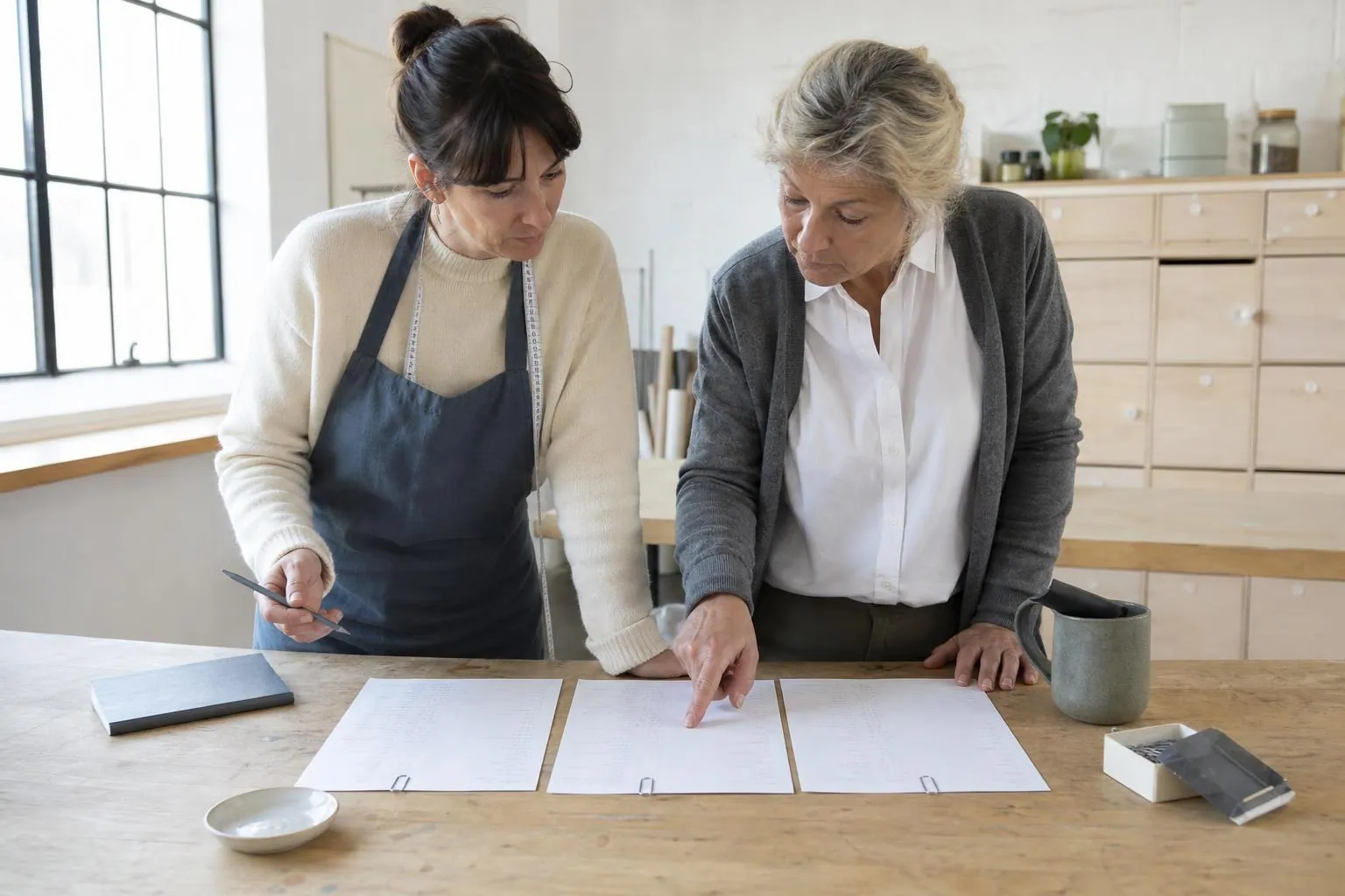 Deux artisanes comparant trois feuilles de calcul étalées sur une table d’atelier partagé, une pièce de céramique posée à côté