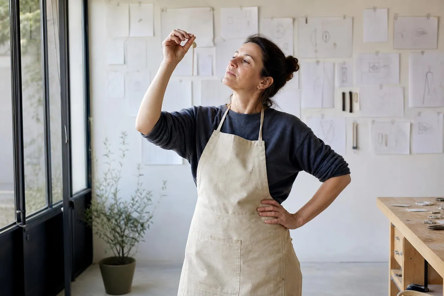 Créatrice de bijoux dans son atelier, face à un mur clair où sont épinglées des fiches de gamme tenues par des trombones